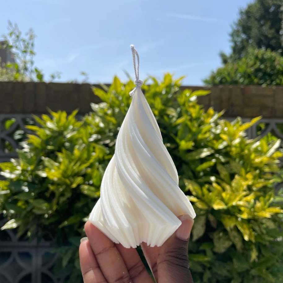 Hand-poured white soy pillar candle with twisted design from The Twirl collection held outdoors against clear sky and green foliage background