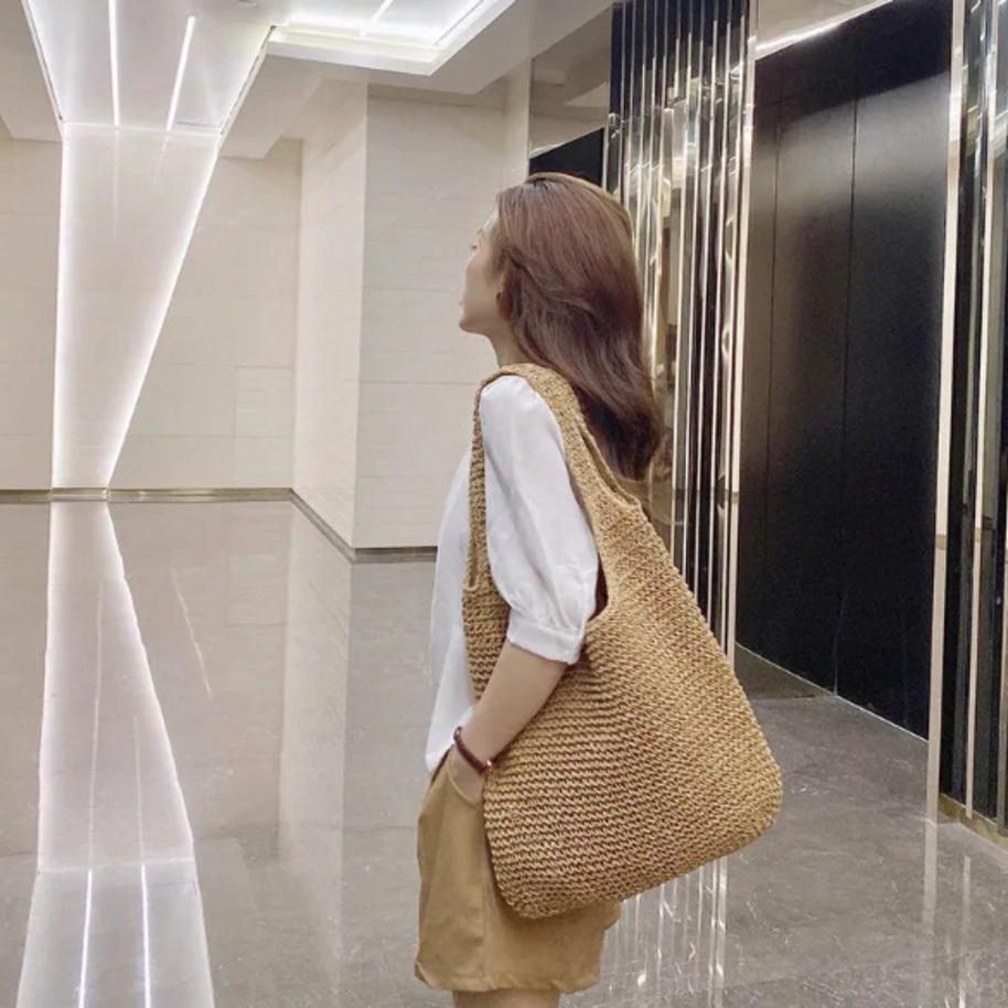Casual summer shoulder beach bag made of straw with a spacious interior, worn by a woman in a white blouse and tan shorts in a modern indoor setting