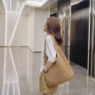 Casual summer shoulder beach bag made of straw with a spacious interior, worn by a woman in a white blouse and tan shorts in a modern indoor setting