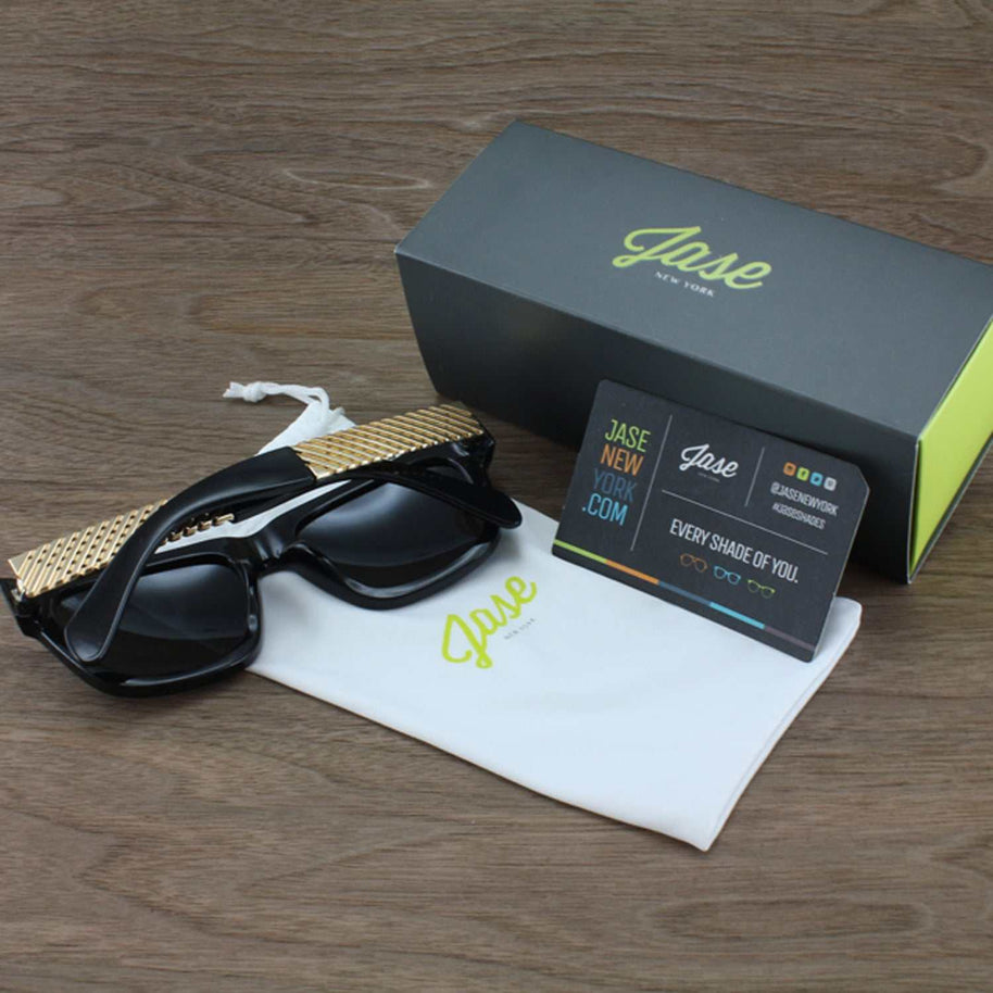 Jase New York Royce sunglasses in gloss black with rectangular frame and metal cross hatch temples displayed with branded box and cloth on wooden surface